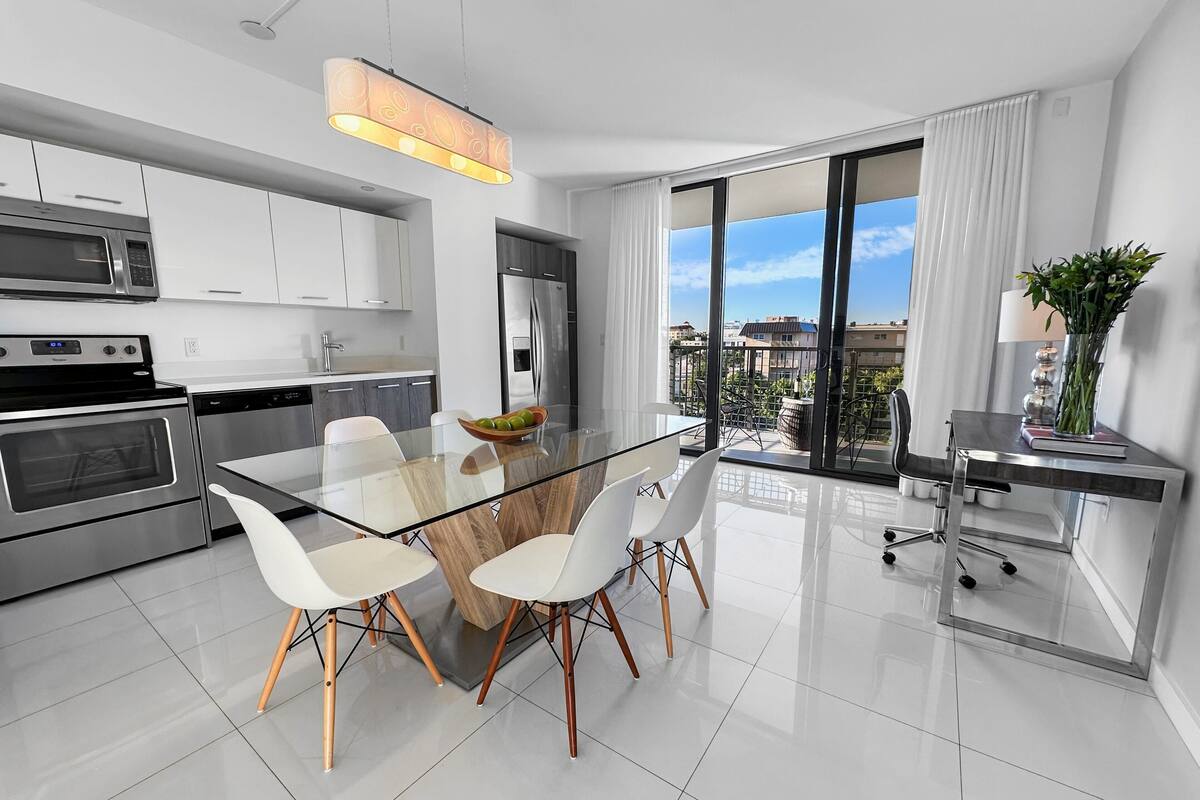 A modern kitchen with stainless steel appliances, a glass dining table, and a view of the city through large windows.