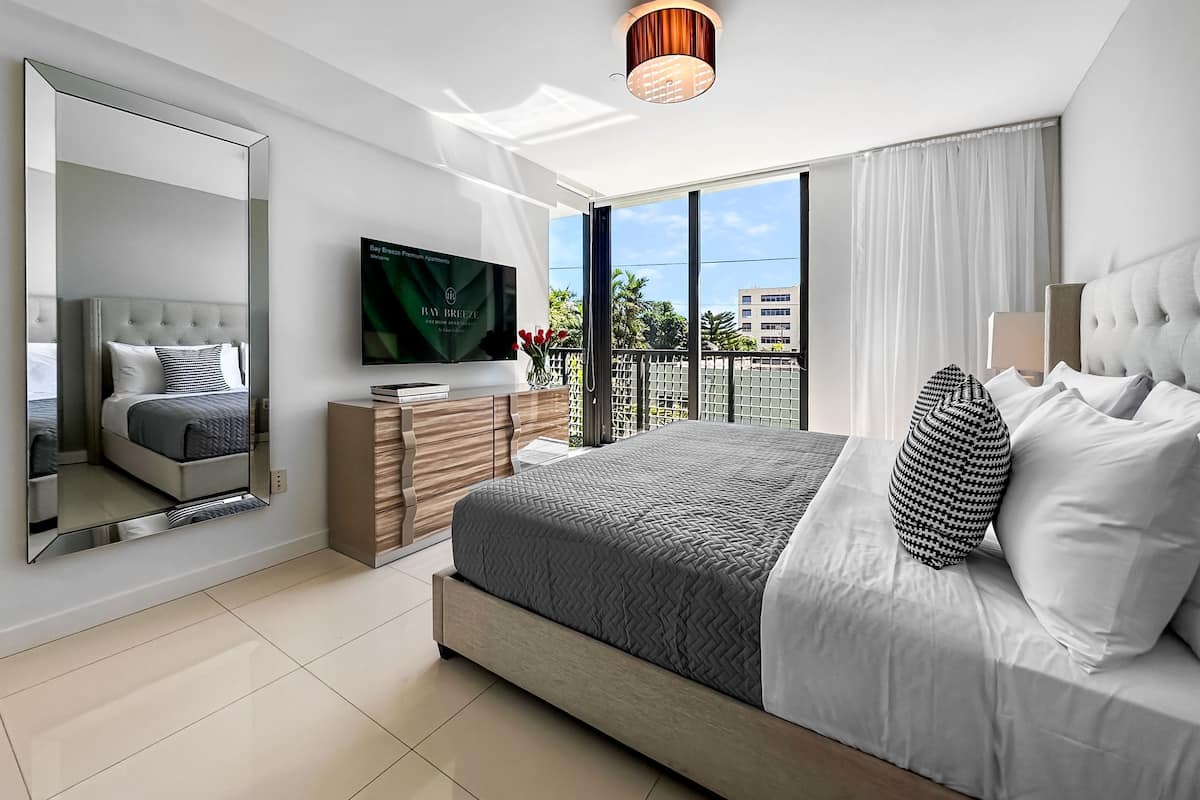 A modern bedroom with a large bed, a TV, a dresser, and a balcony with a view.