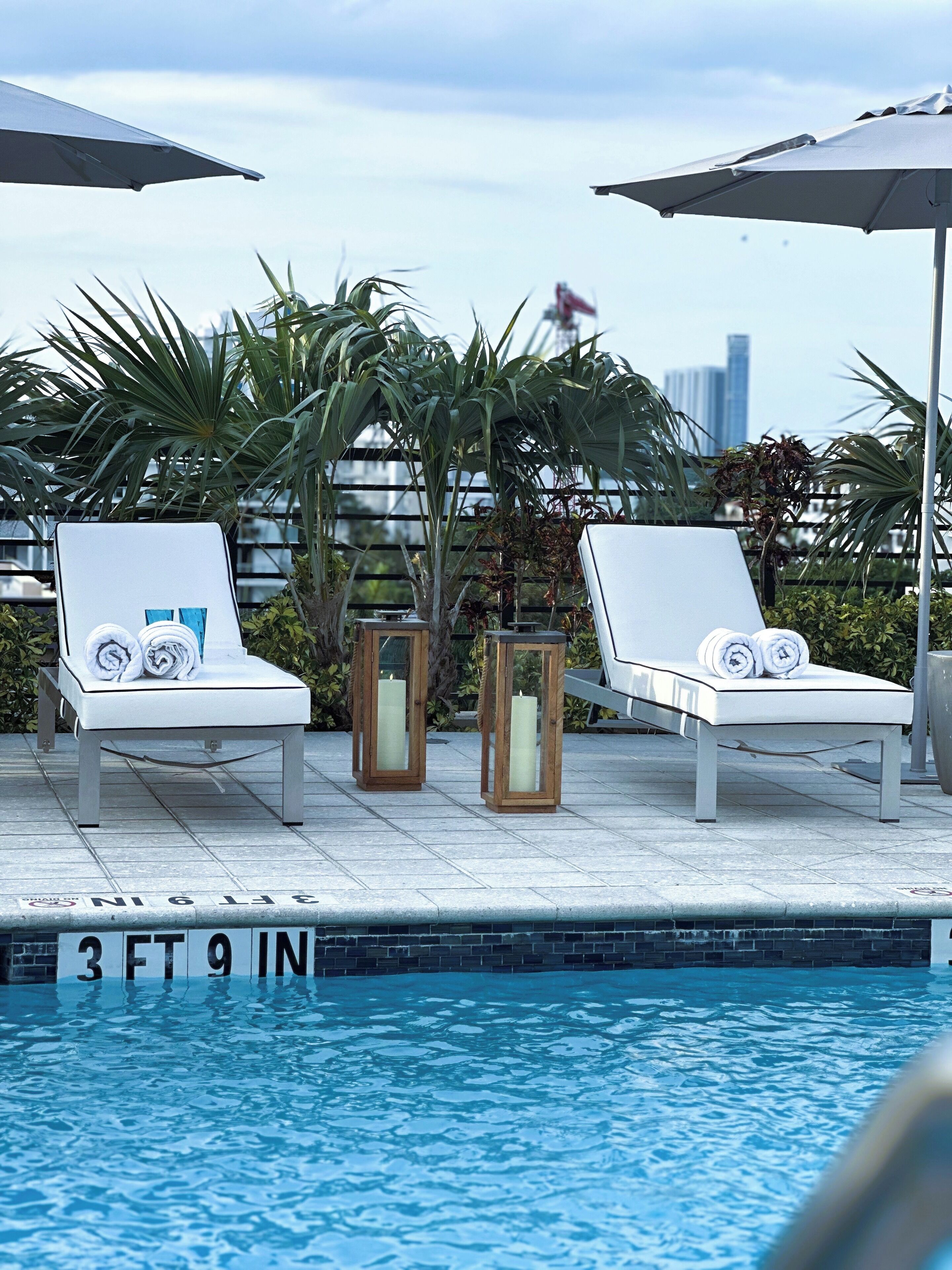 A poolside area with two lounge chairs, a sign indicating a 3ft 9in pool depth, and a city skyline in the background.