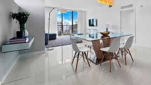 A modern living room with a glass dining table, white chairs, a wooden coffee table, a floor lamp, and a view of the city through large windows. - Bay Breeze Premium Apartments By Eskape Collection (Bay Harbor Islands)