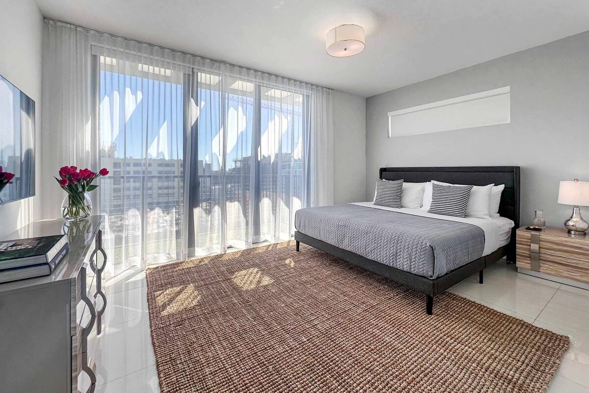 A modern bedroom with a large bed, a bedside table, a nightstand, and a view of the city through sheer curtains.