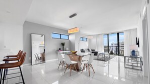 A modern, well-lit living room with a large window, a dining area with a glass table, and a sofa. - Bay Breeze Premium Apartments By Eskape Collection (Bay Harbor Islands)