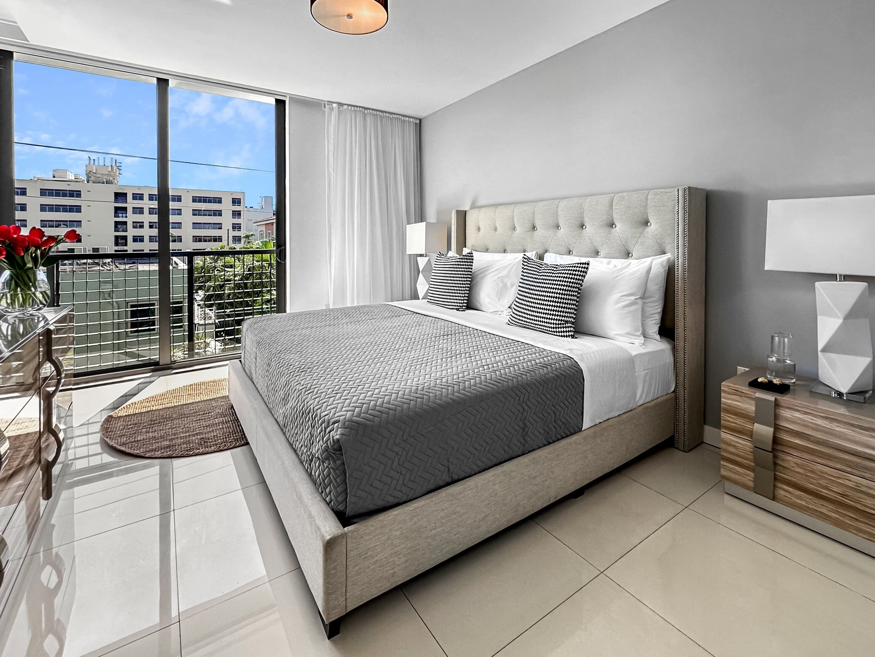 A modern bedroom with a large bed, a nightstand, a lamp, and a view of buildings through a balcony.
