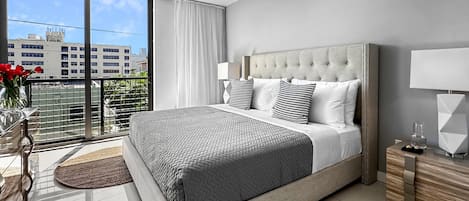 A modern bedroom with a large bed, a nightstand, a lamp, and a view of buildings through a balcony.