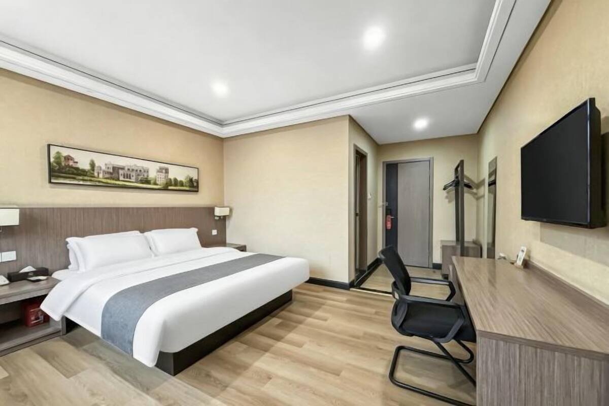 A hotel room with a bed, desk, chair, TV, and artwork on the wall.