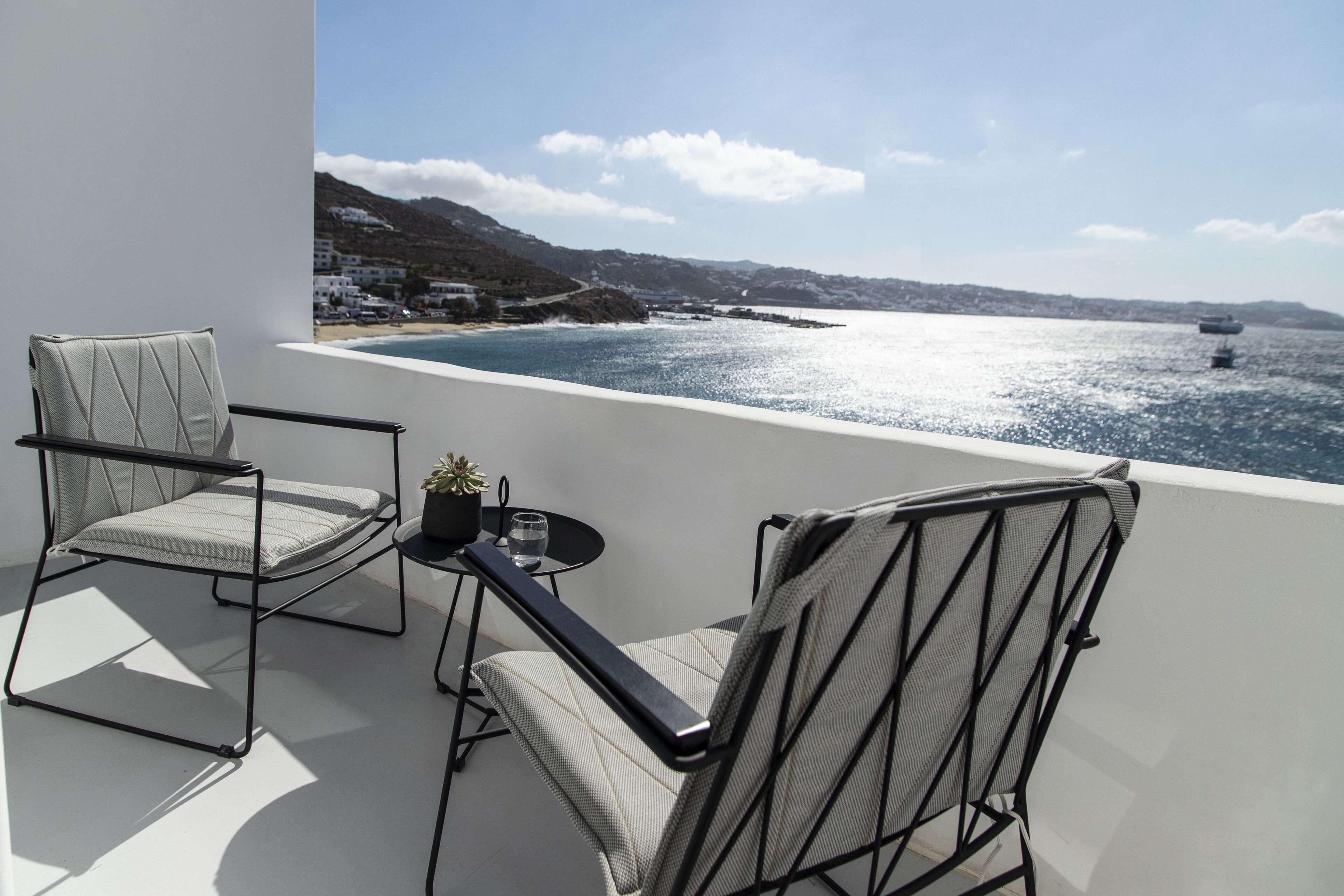 A balcony with two chairs and a small table, overlooking a coastal view with boats and buildings.