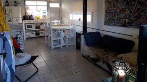 Ein geräumiger Wohnbereich mit Esstisch, weißen Stühlen, einer Couch und einem Fernseher. Es gibt eine Küchenzeile mit Geräten und ein Fenster.