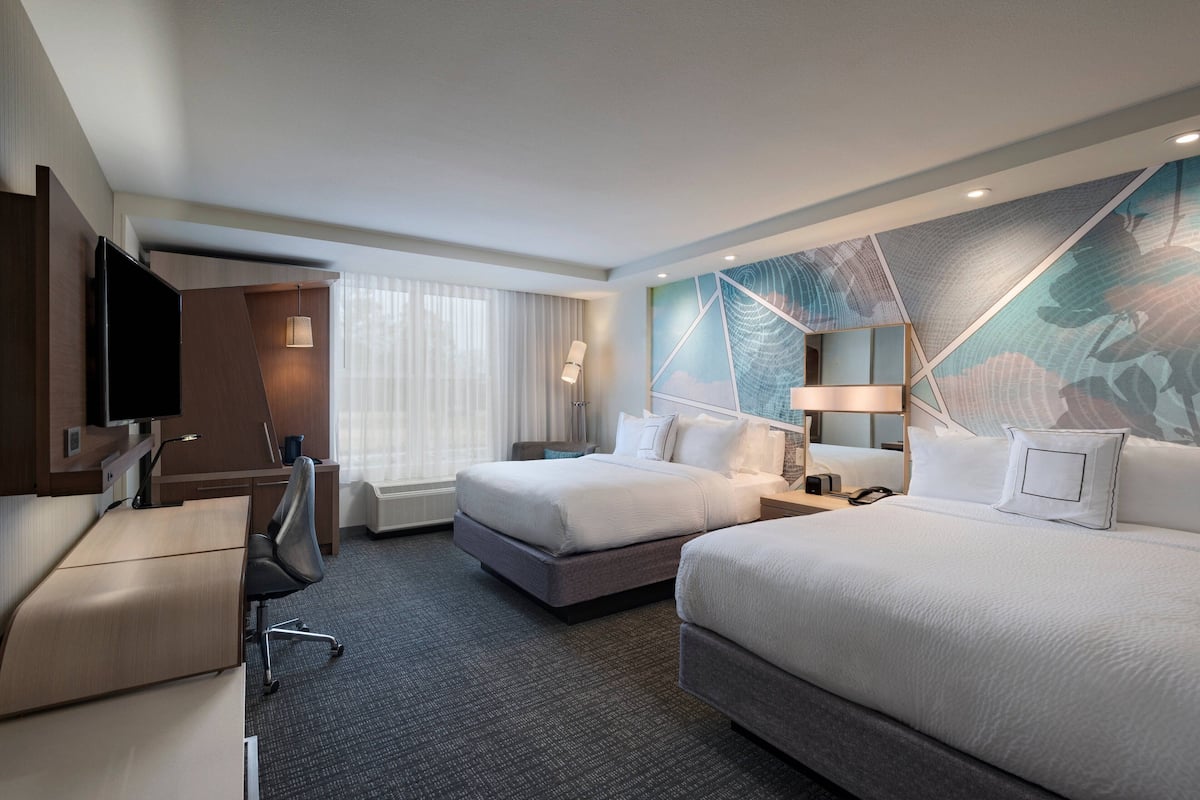 A hotel room with a desk, two beds, a TV, and a large window with curtains.