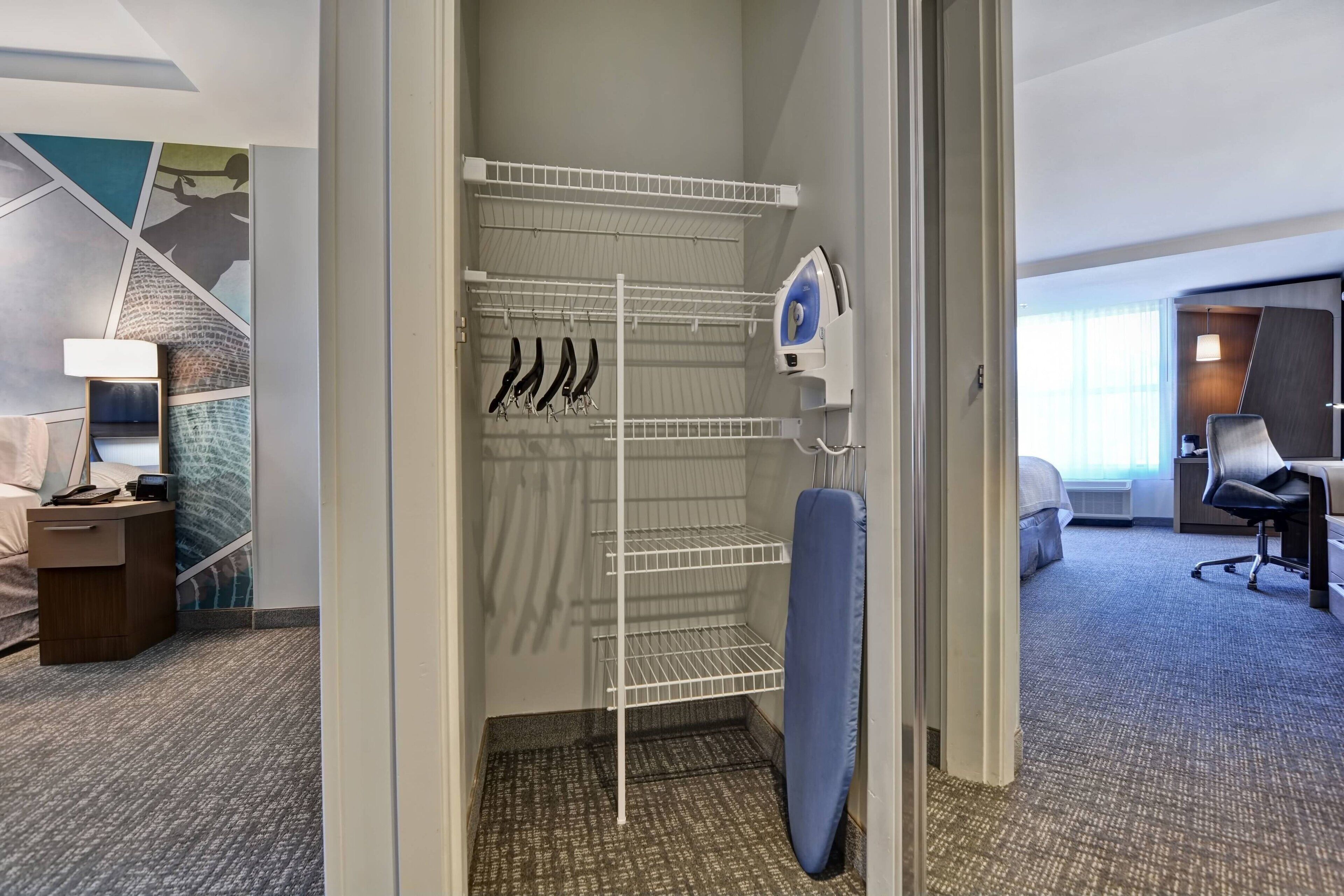 Habitación de hotel con cama, escritorio, silla, armario con ropa colgada y una tabla de planchar azul.