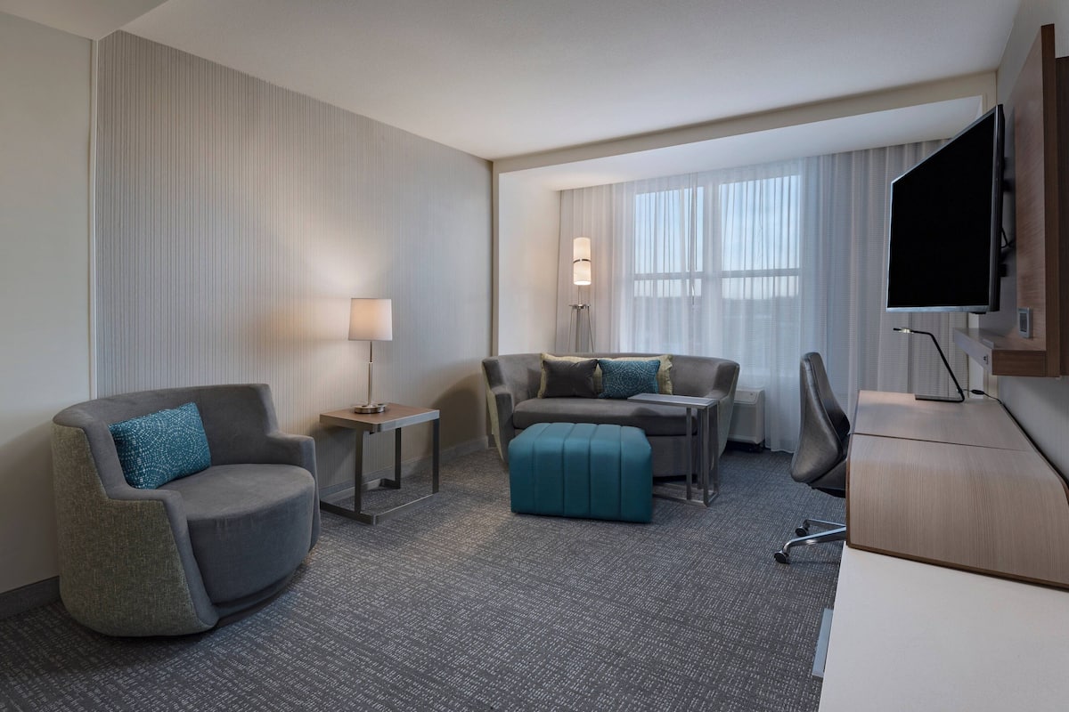 A hotel room with a grey armchair, a sofa with a teal ottoman, a desk with a flat-screen TV, and a large window with sheer curtains.