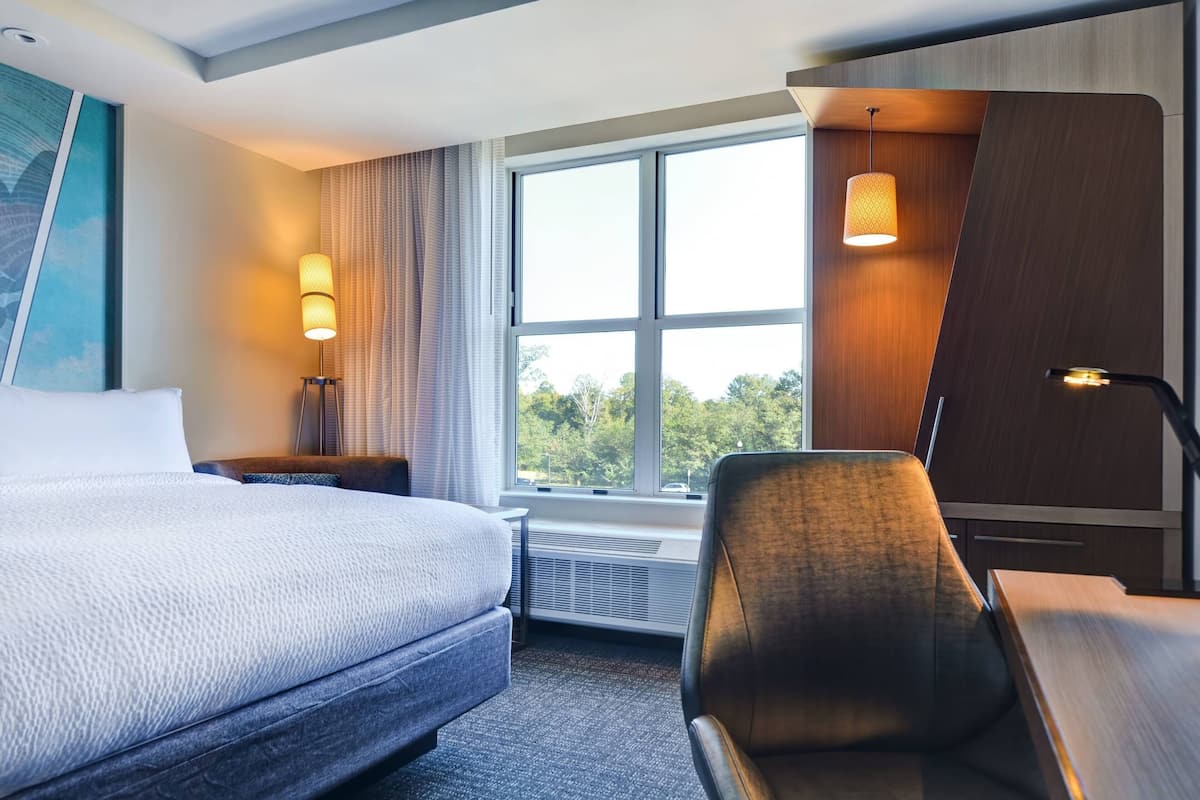 A hotel room with a large bed, a desk, a chair, a window with a view of trees, and a painting on the wall.