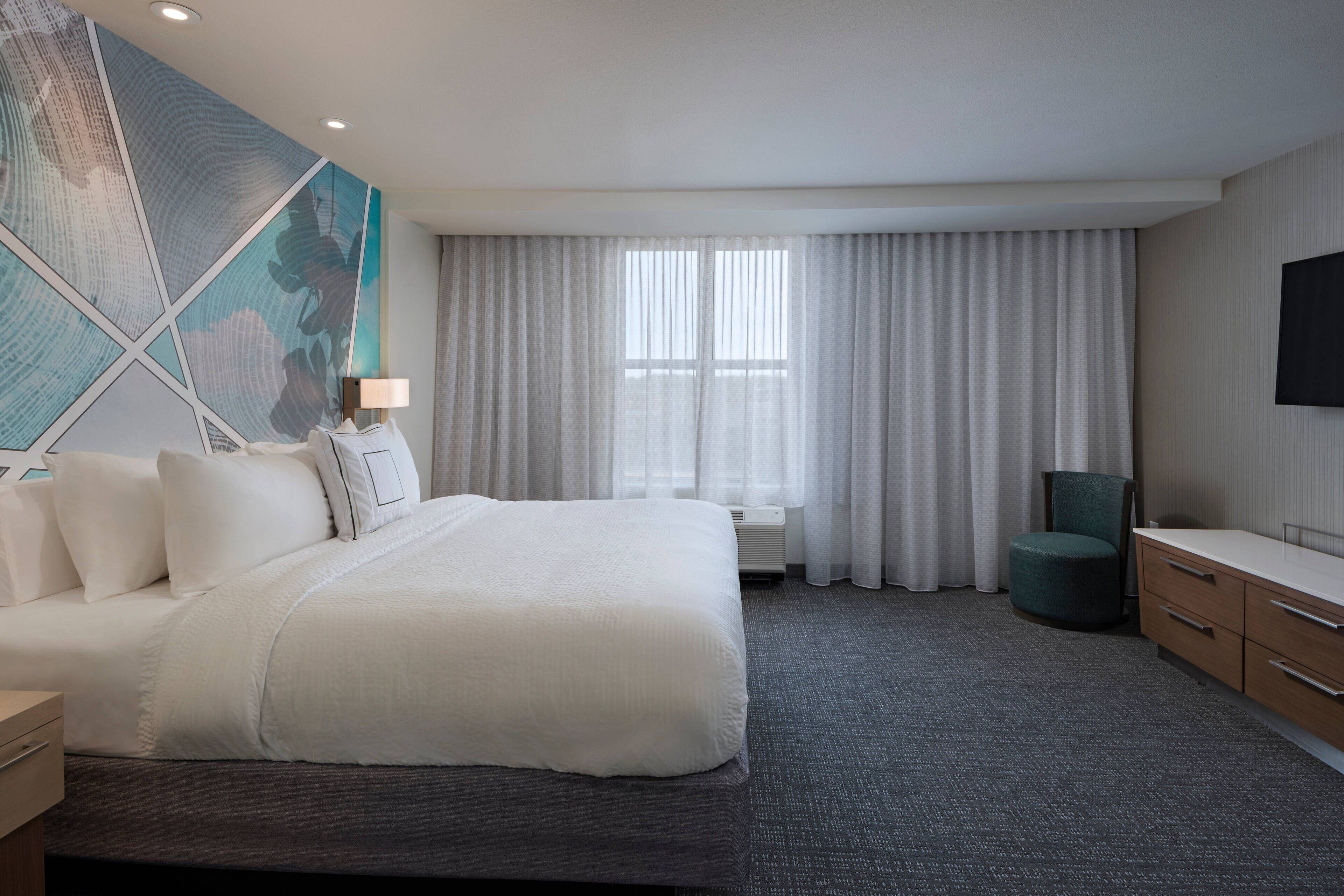 A hotel room with a large bed, a desk, a chair, a flat-screen TV, and a large window with curtains.