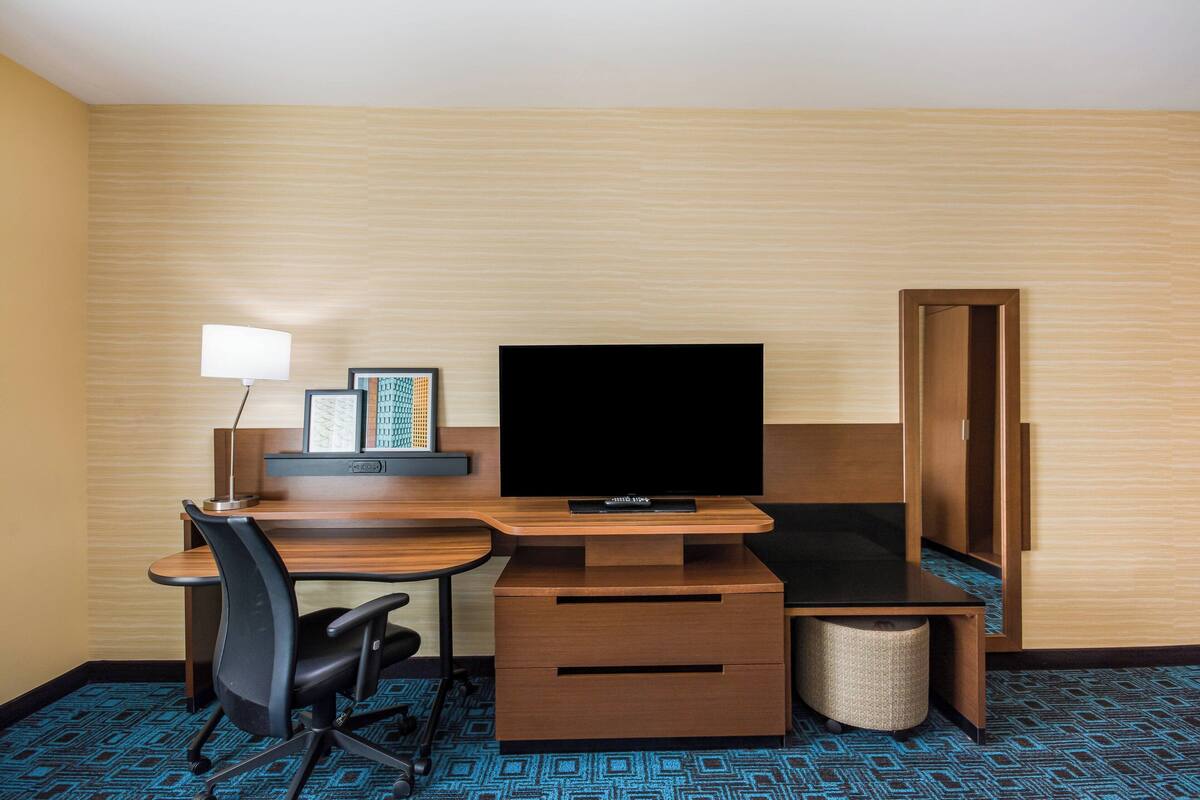 A hotel room with a wooden desk, a black office chair, a television, a lamp, a waste basket, and a framed picture on the wall.