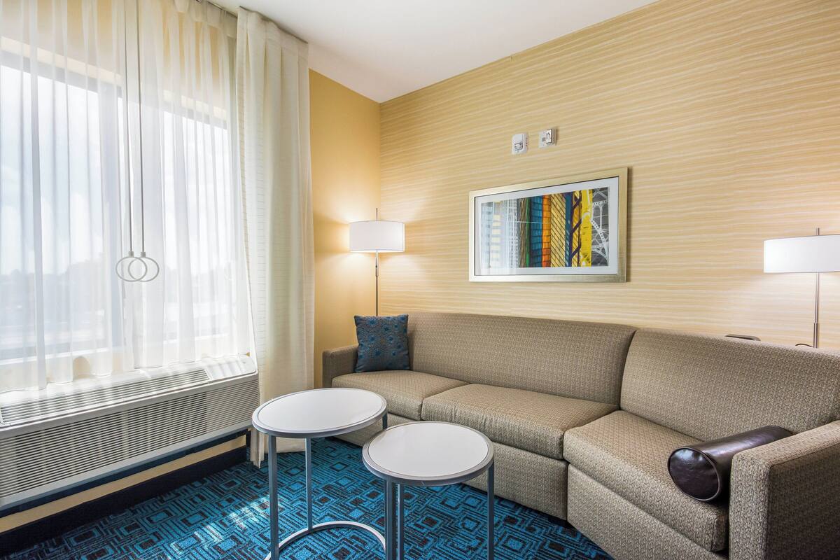 A hotel room with a beige sofa, two round tables, a large window with sheer curtains, a radiator, a wall-mounted painting, and two floor lamps.