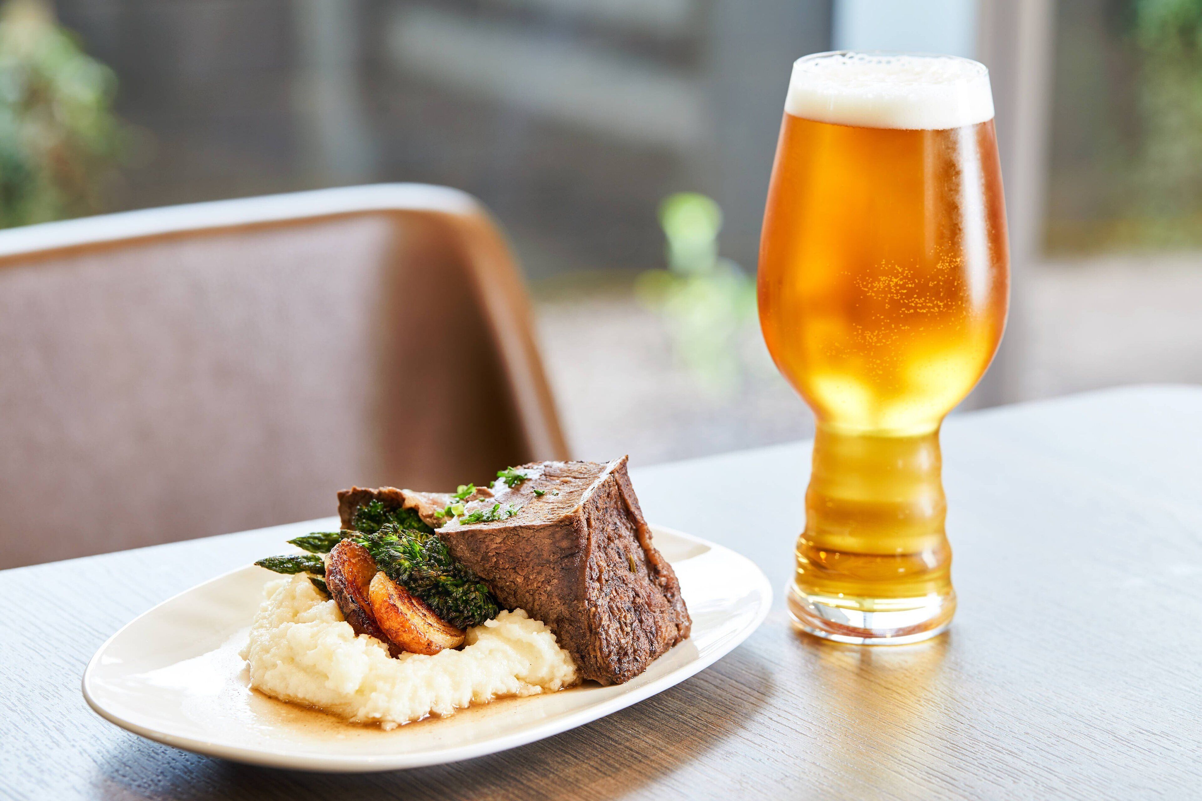 Un plato de comida con puré de patatas, carne asada y verduras, junto con una jarra de cerveza sobre una mesa de madera.