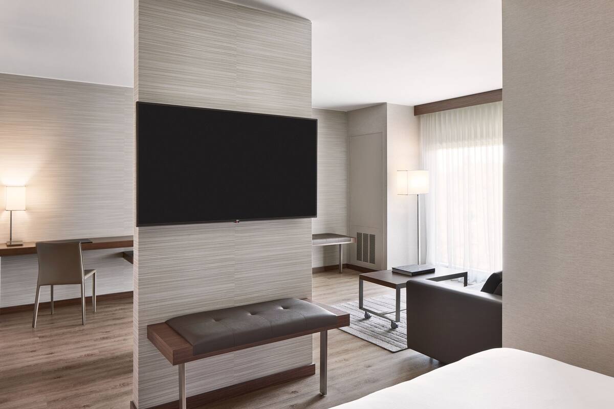 A modern hotel room with a flat-screen TV mounted on the wall, a small desk with a chair, a bench, a sofa, and a window with curtains.
