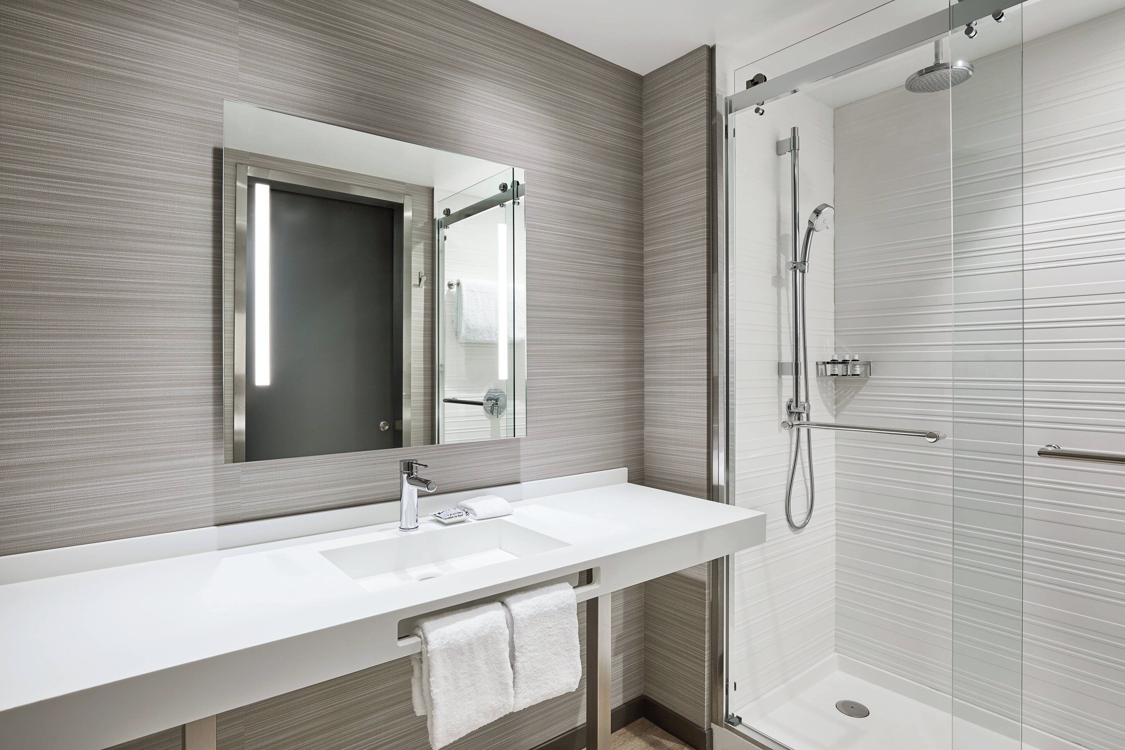 Un baño moderno con un espejo grande, un lavamanos blanco y una ducha con mampara de cristal.