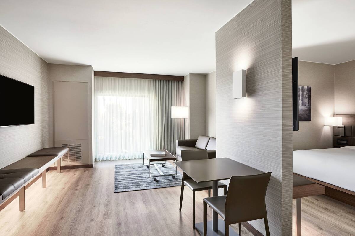 A modern hotel room with a flat-screen TV, a sofa, a dining table with chairs, a bed, and a large window with curtains.