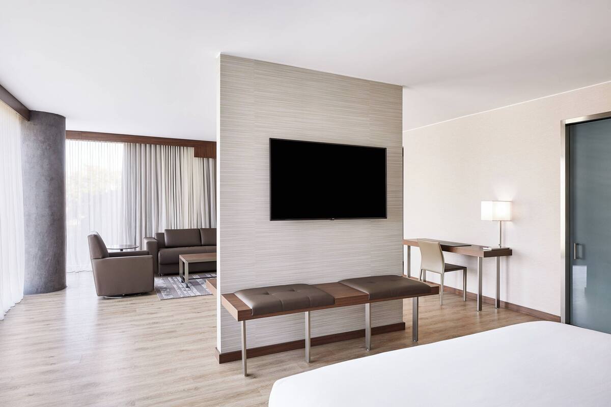 A modern hotel room with a large bed, a flat-screen TV mounted on the wall, a seating area with a sofa and chairs, and a desk with a lamp.