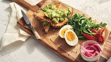 A wooden cutting board with toast topped with avocado, a halved boiled egg, arugula, and pickled red onions.