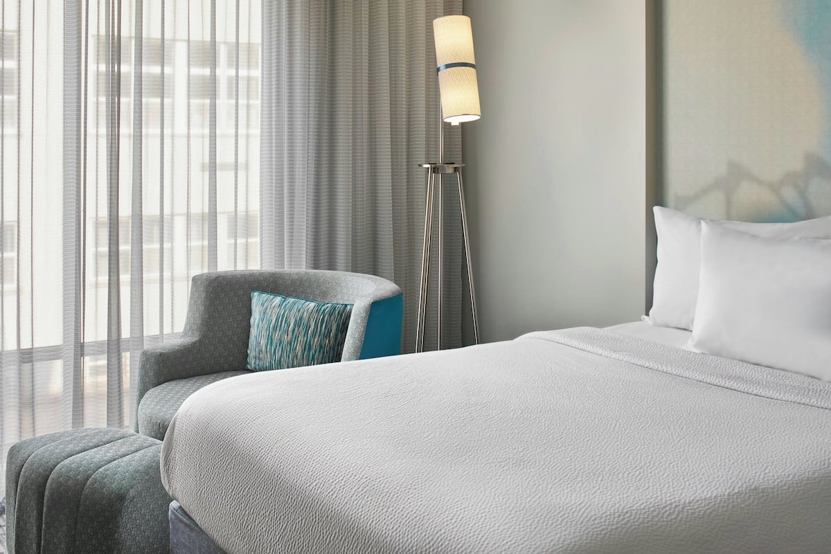 Ein Hotelzimmer mit einem großen Bett, einem Sessel mit Kopfstütze, einer Stehlampe und Blick auf die Stadt durch transparente Vorhänge.