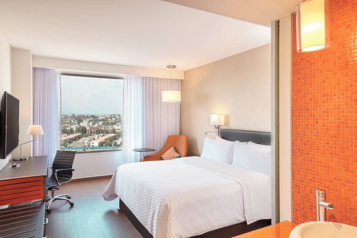 A hotel room with a bed, a desk with a TV, a chair, a window with a view, and a bathroom with a sink and mirror.