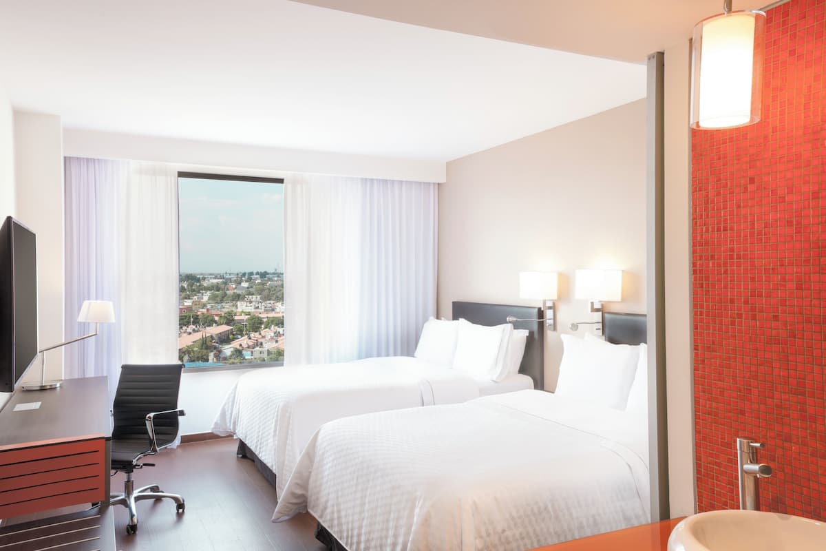A hotel room with two beds, a desk, a chair, a TV, and a bathroom with a red tiled wall.