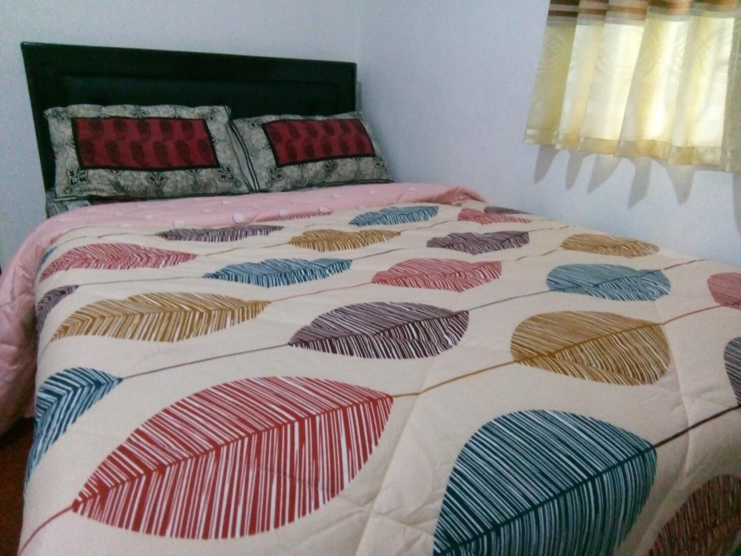 Un lit avec une couette à motifs et deux oreillers, une tête de lit sombre et une fenêtre avec un rideau léger.