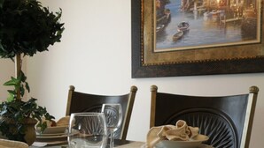 Una mesa de comedor con cubierta de vidrio, dos sillas, un plato con comida, un vaso y un cuadro enmarcado en la pared.