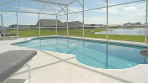 Una piscina con zona techada, rodeada por una valla blanca, y vista a una casa y paisaje al fondo.