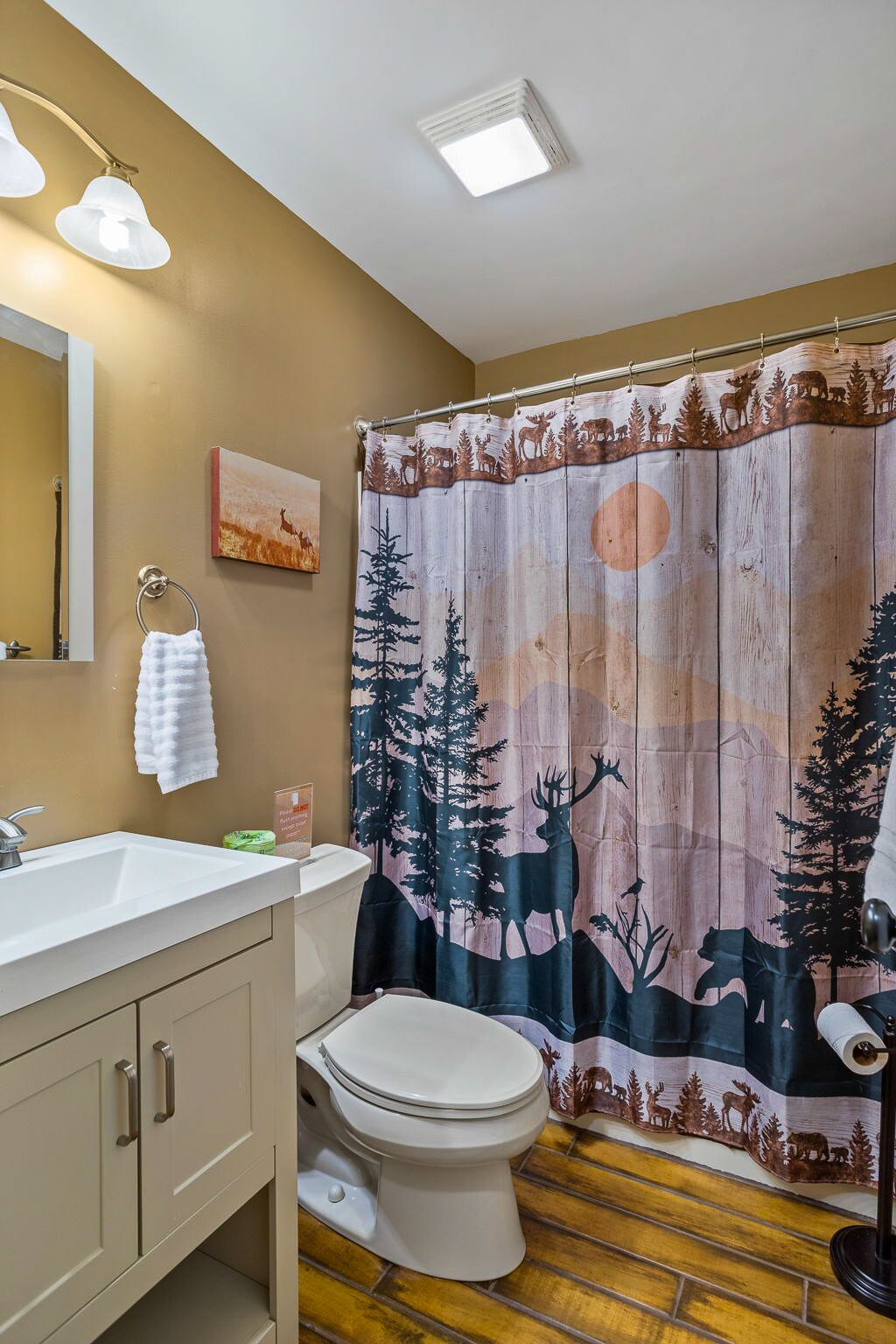 Ein Badezimmer mit Holzboden, einem weißen Waschbecken, einer Toilette und einer Duschvorhang mit Wald- und Tierwelt-Motiv.