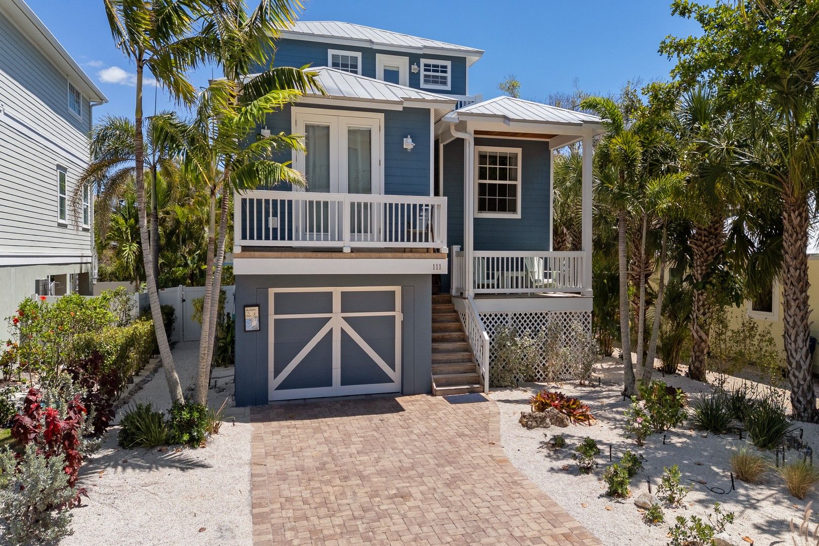 A two-story house with a covered front porch, a garage, and a landscaped garden with palm trees and shrubs.