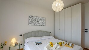 A neatly made bed with a white comforter, a bedside lamp, a glass bedside table with a lamp, a framed floral artwork, and a tray with towels and a flower on the bed.