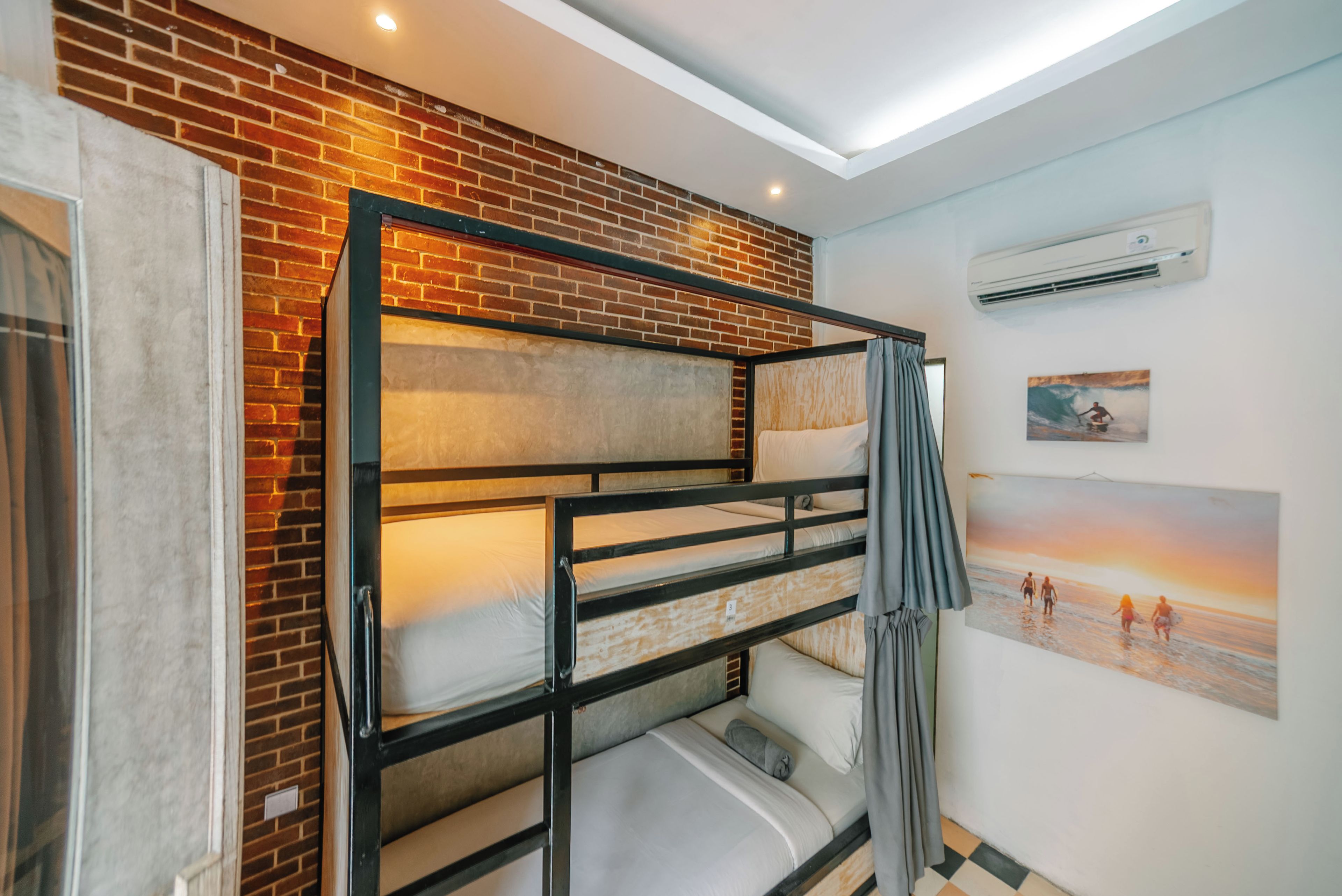 Une chambre avec un lit superposé, un mur en briques, un climatiseur et un tableau représentant une scène de plage.