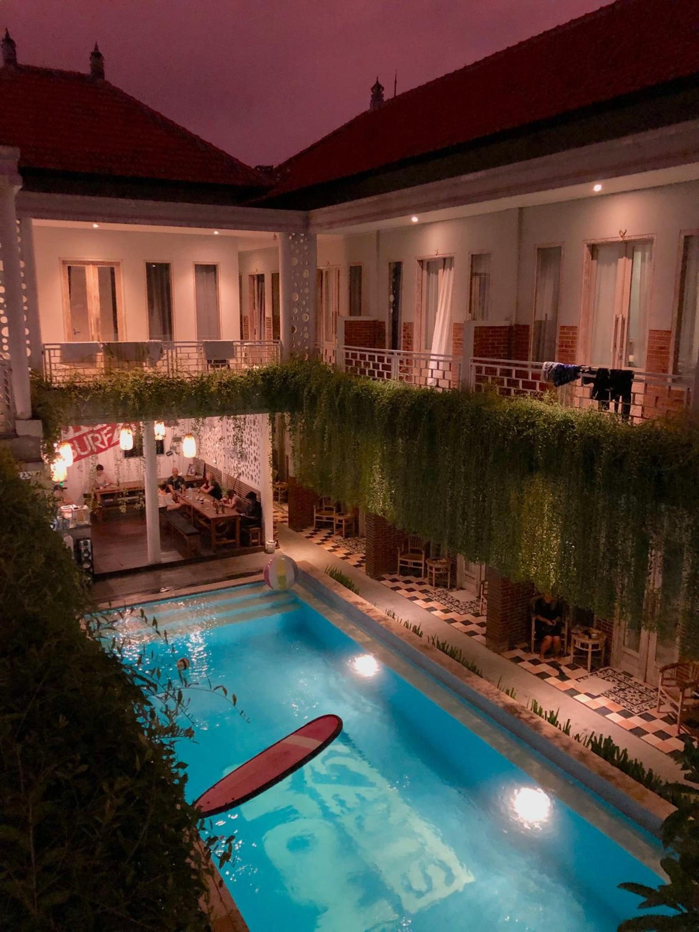 Zona de piscina con suelo a cuadros, un área de descanso con mesas y sillas, y un edificio con múltiples ventanas y balcones.