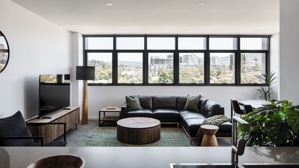 A modern living room with a large window, a black leather sofa, a round coffee table, and a flat-screen TV. - Astra Apartments Wollongong (Wollongong)