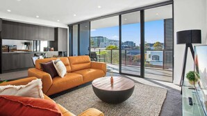 A modern living room with a large sofa, a coffee table, and a view of the city through large windows. - Astra Apartments Wollongong (Wollongong)