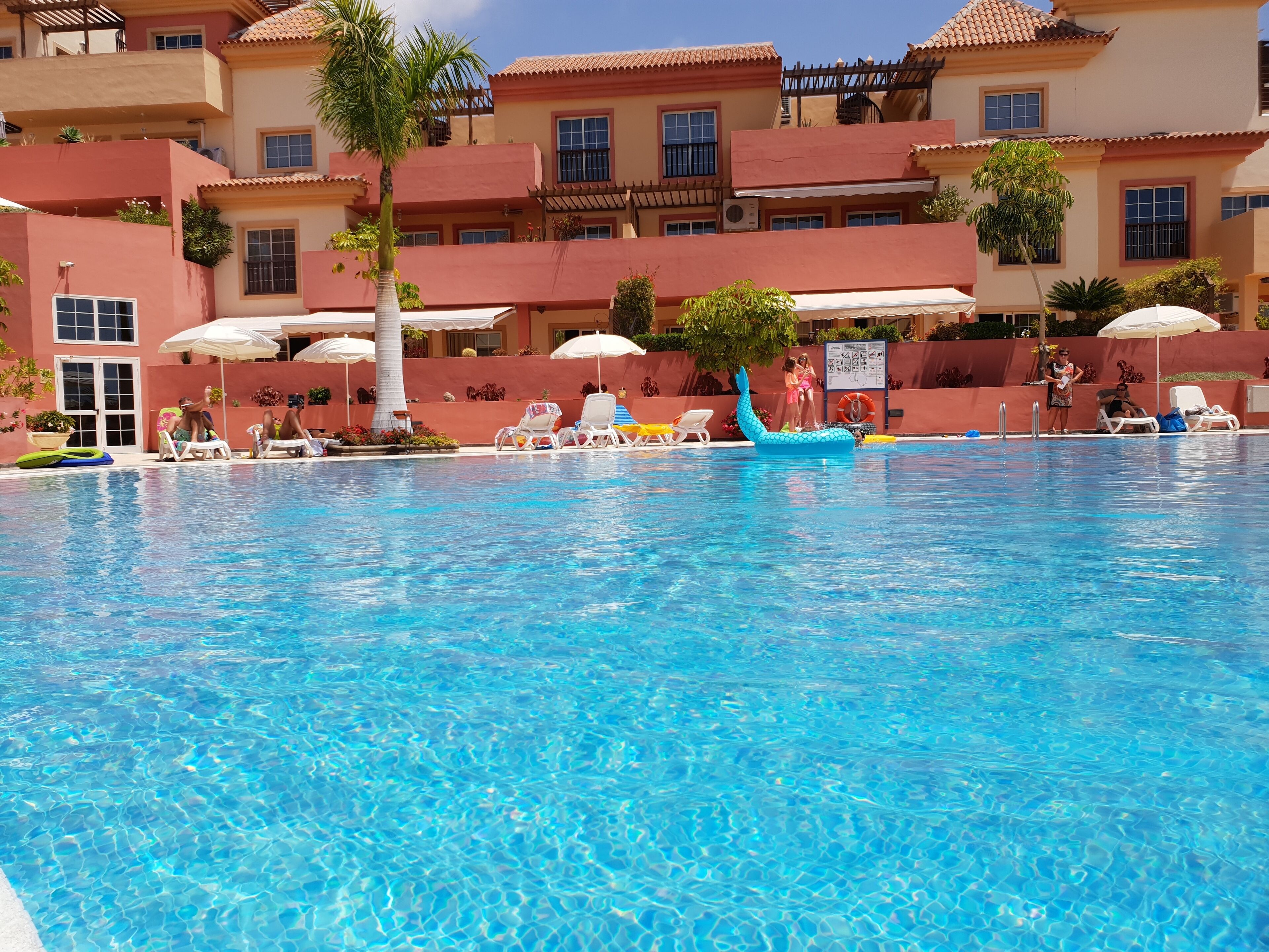 Ein Schwimmbecken mit Liegestühlen und Sonnenschirmen, umgeben von mehrstöckigen Gebäuden mit Balkonen und Fenstern.
