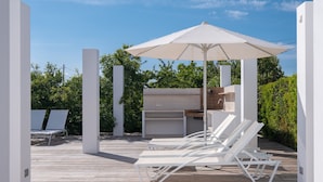 Une terrasse avec des chaises longues blanches, un barbecue et un grand parasol.