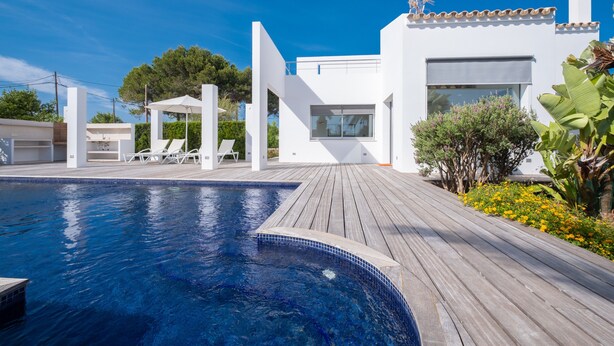Un espace piscine moderne avec une terrasse en bois, un bâtiment blanc doté de grandes fenêtres et un ciel d’un bleu éclatant.