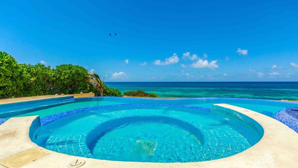 A circular infinity pool with clear blue water, surrounded by a concrete deck and lush greenery, overlooking a turquoise ocean.
