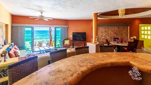 A spacious living area with a large window overlooking the ocean, a ceiling fan, and a dining area with a round table and chairs.