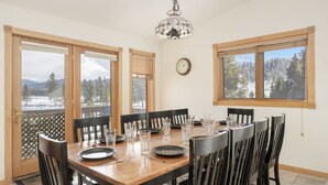 A dining area with a wooden table set for six, surrounded by black chairs. There are three windows providing views of a snowy landscape. - Mountain Views w/ Hot Tub (Tabernash)
