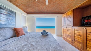 A bedroom with a large bed, a ceiling fan, a wooden wardrobe, and a view of the sea.