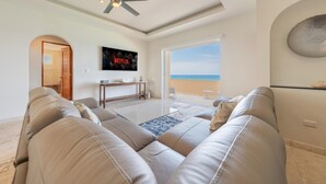 A modern living room with a large sofa, a coffee table, and a TV mounted on the wall. There is a ceiling fan, a door leading to another room, and a view of the beach through a large window.