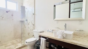 A modern bathroom with a glass-enclosed shower, a white toilet, a sink with a marble countertop, and a large mirror above the sink. - Modern 1 BR + Pool. Heart of Downtown (Isla Mujeres)