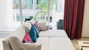 A modern living room with a beige sectional sofa, colorful throw pillows, and a large window offering a view of the outdoors. - Modern 1 BR + Pool. Heart of Downtown (Isla Mujeres)