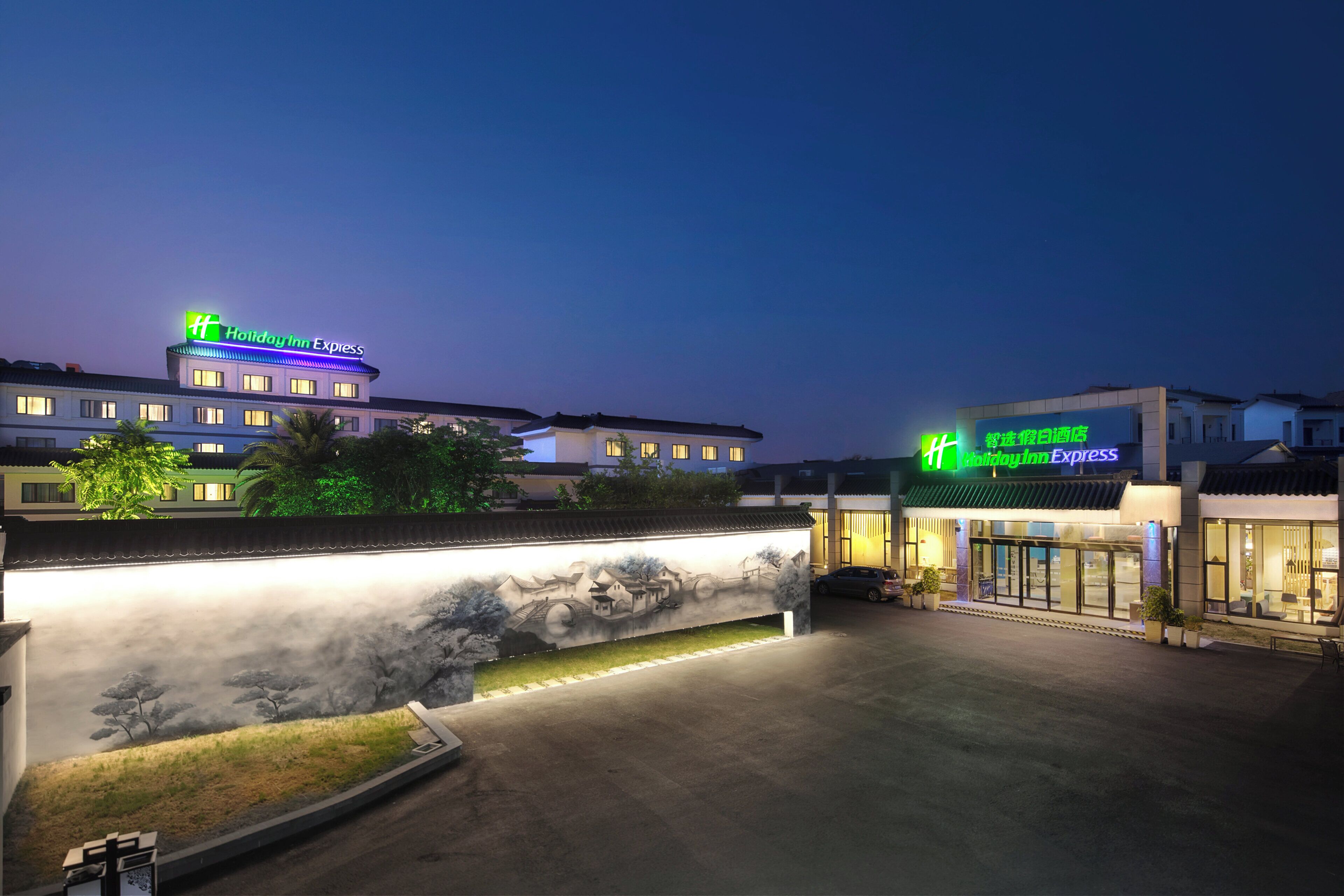 A Holiday Inn Express hotel with a traditional Asian architectural design, a landscaped entrance area, and a large mural.