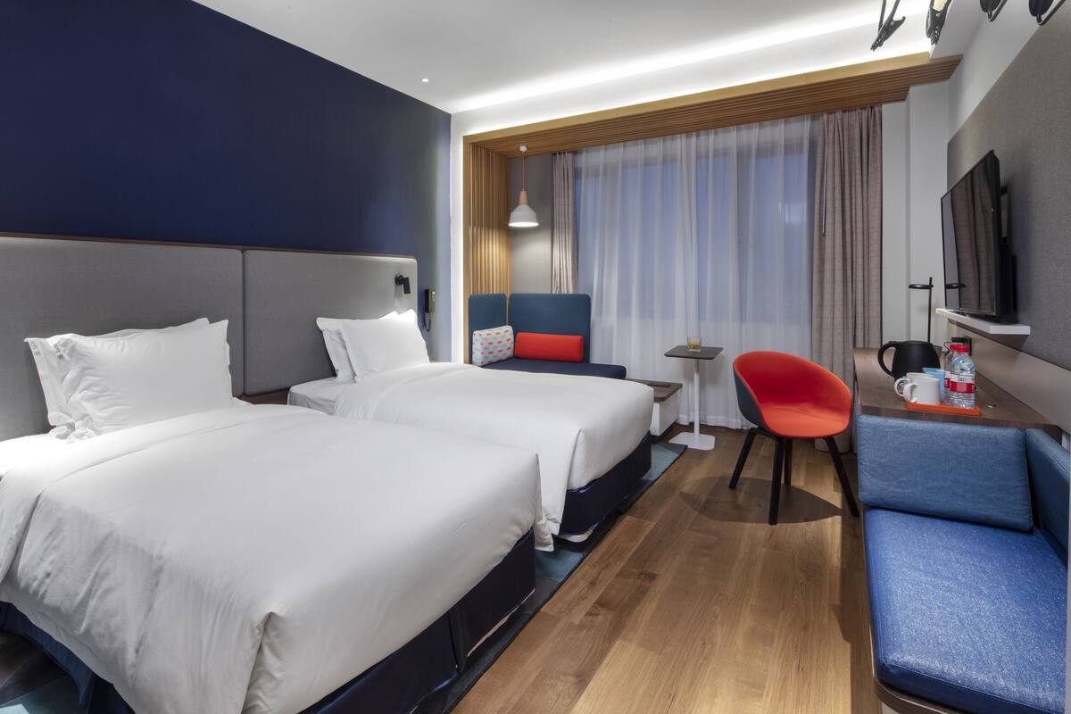 Ein Hotelzimmer mit zwei Betten, einem Schreibtisch, einem Stuhl, einem Fernseher und einem Fenster mit Vorhängen.