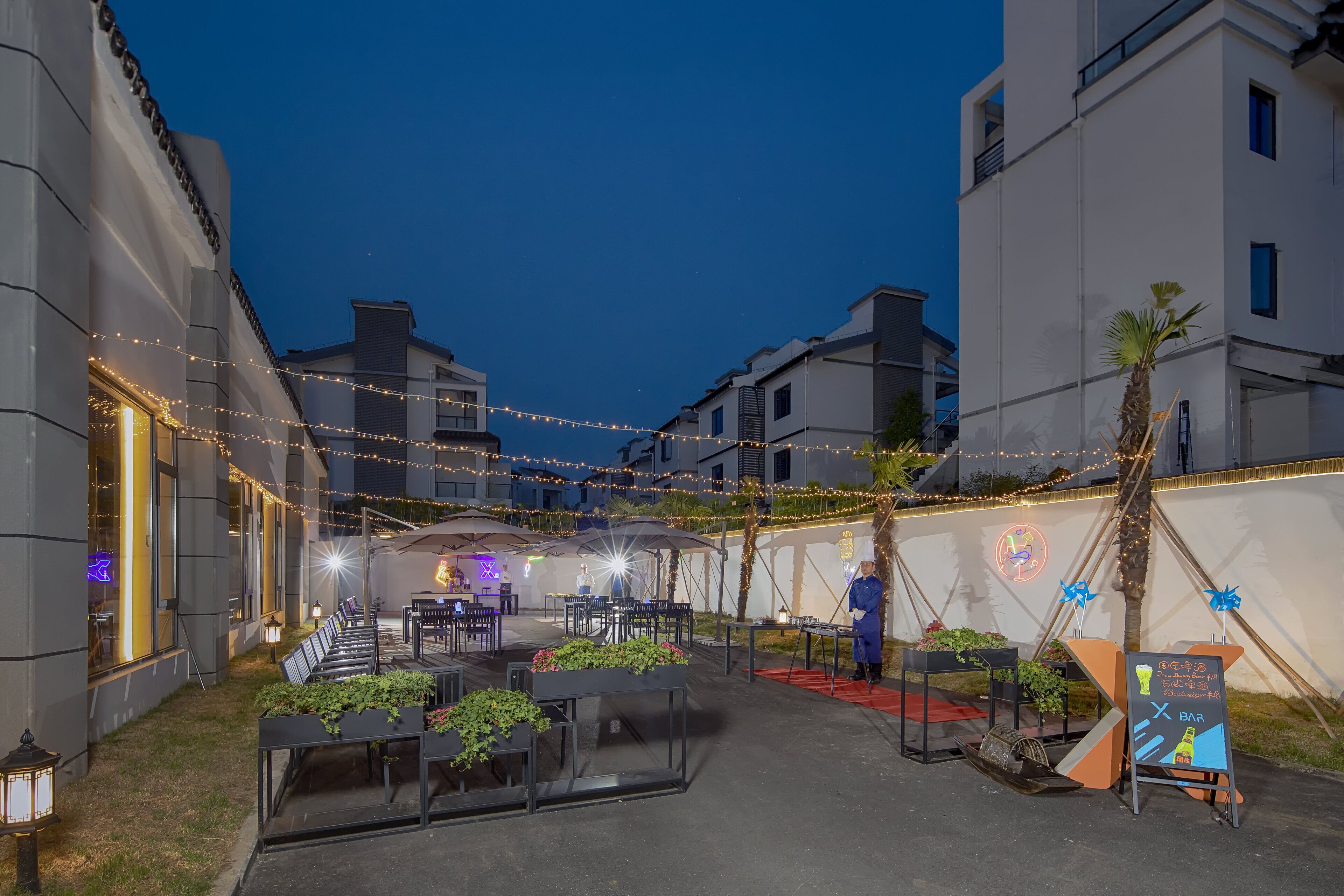 An outdoor seating area with tables and chairs, string lights, and a bar area with a neon sign.